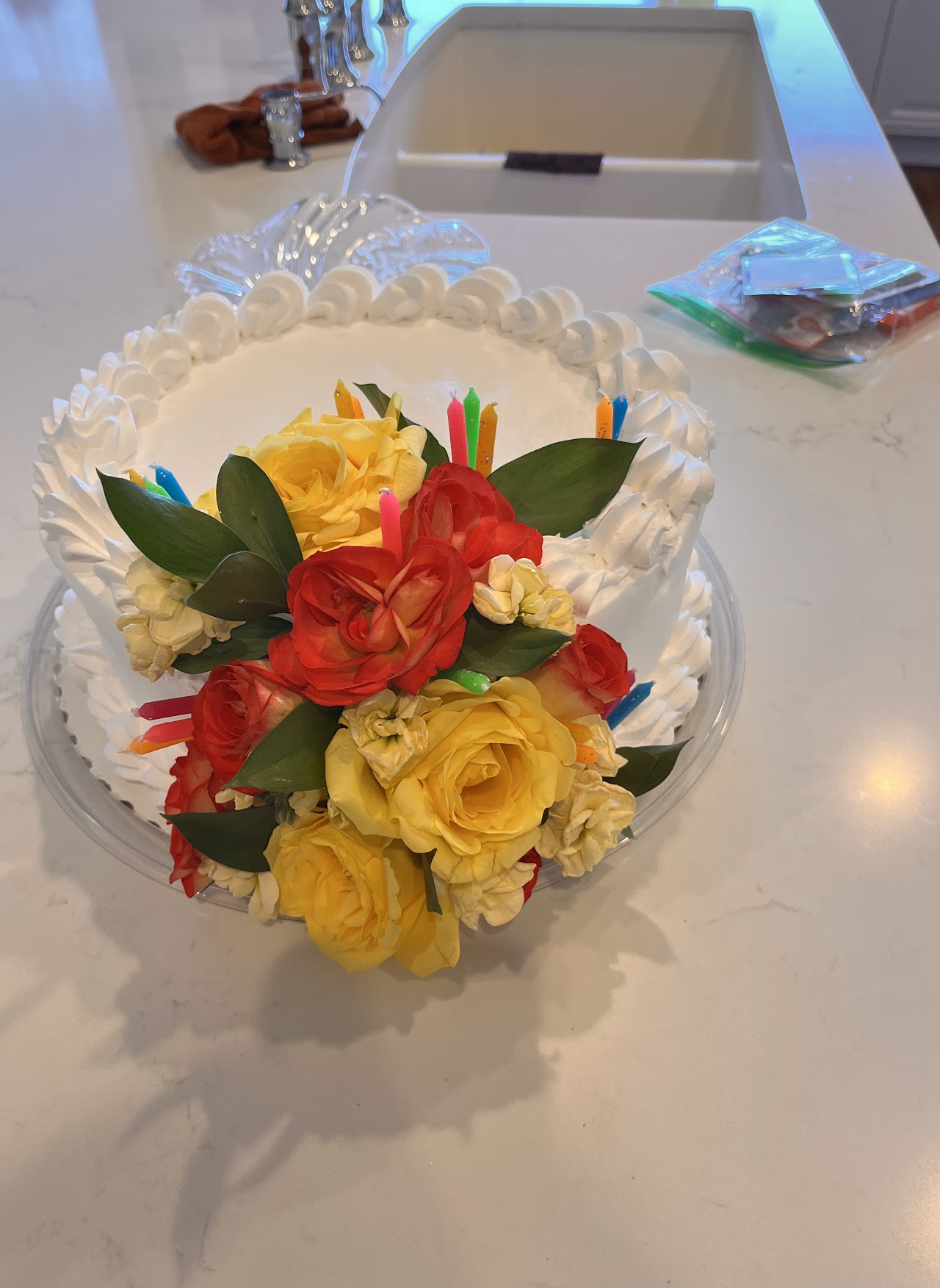 White shell-bordered buttercream cake topped with a fresh arrangement of red and yellow roses and colorful birthday candles