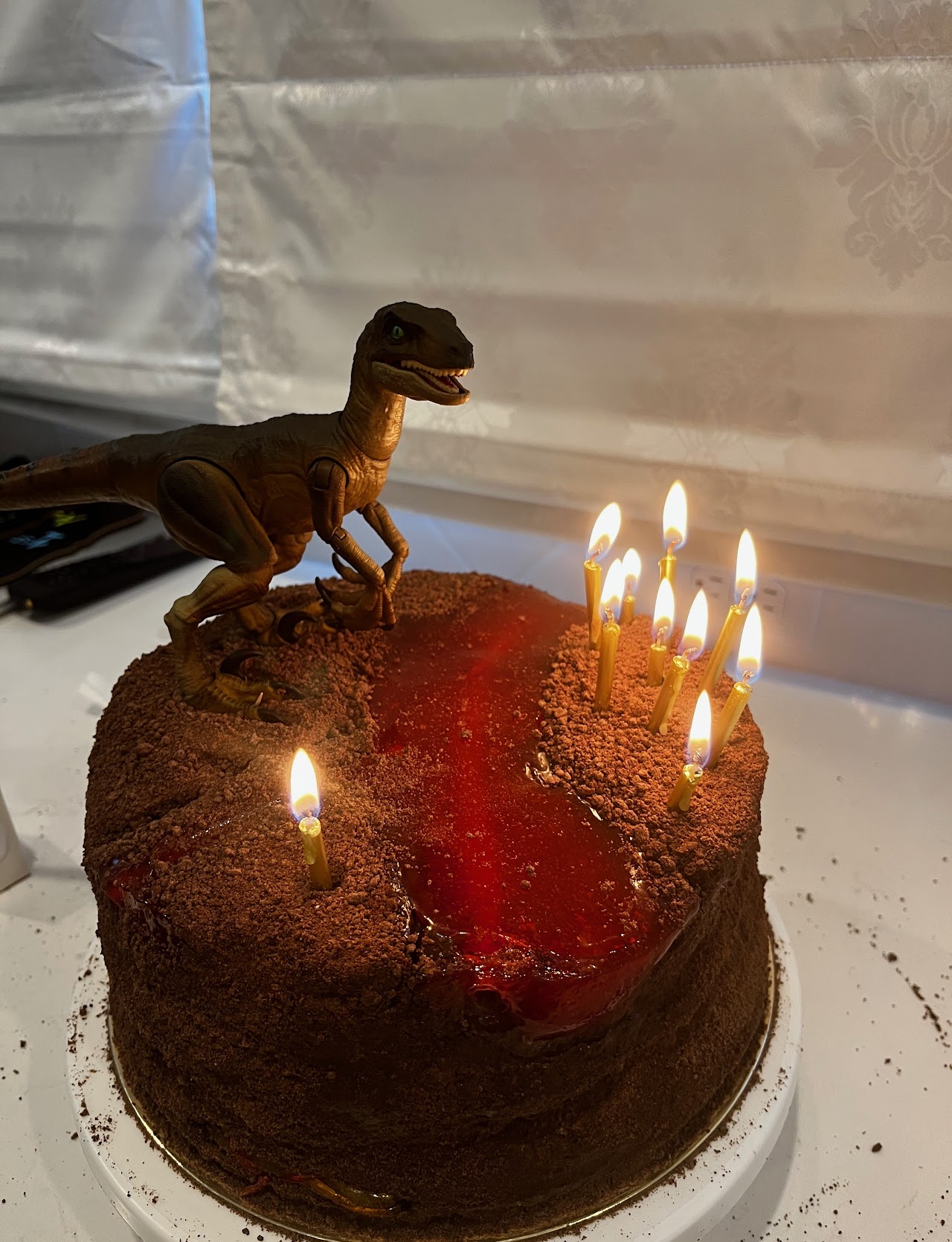 Chocolate cookie crumb covered cake with a red gel lava crack down the center and a raptor figure on top, lit birthday candles
