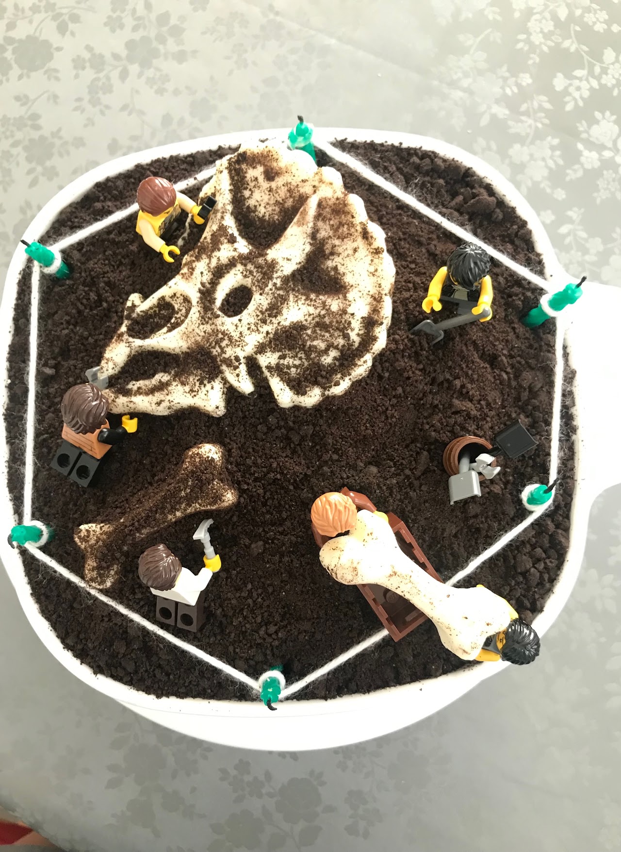 Cookie-crumb dinosaur dig site cake with a piped fondant fossil skull and bone, surrounded by toy paleontologist figures