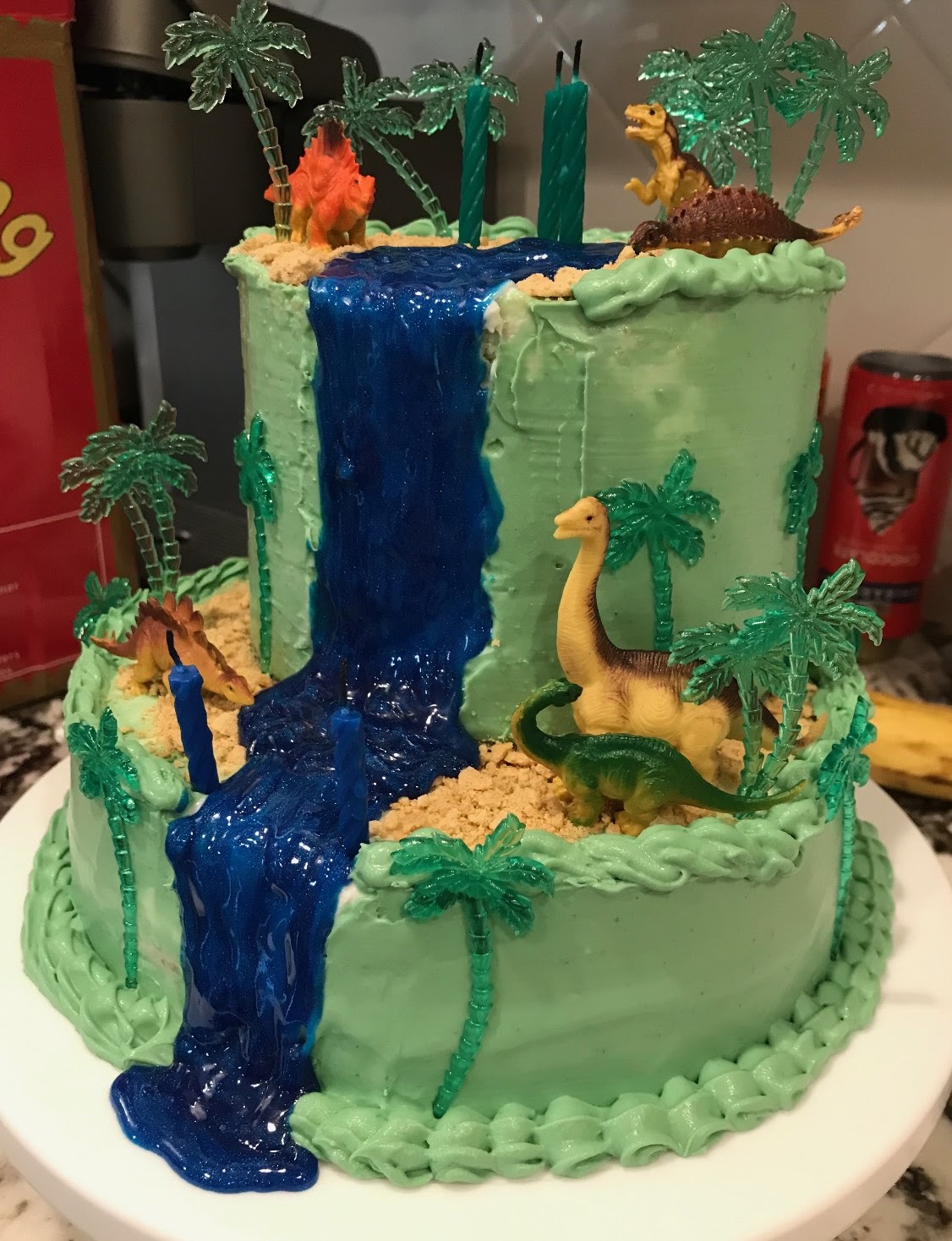 Two-tier mint buttercream cake with a blue gel waterfall, graham cracker riverbank, palm tree picks, and dinosaur figures