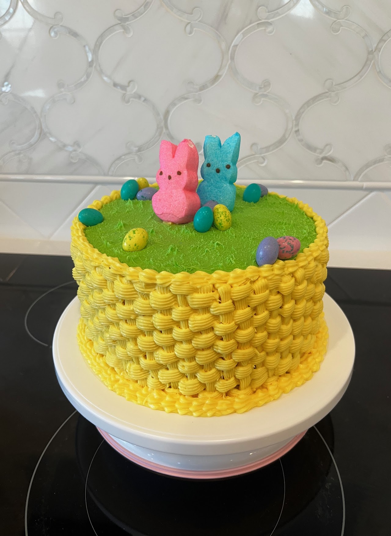 Cake with yellow basket-weave buttercream sides and green grass-textured top, decorated with marshmallow bunnies and candy eggs