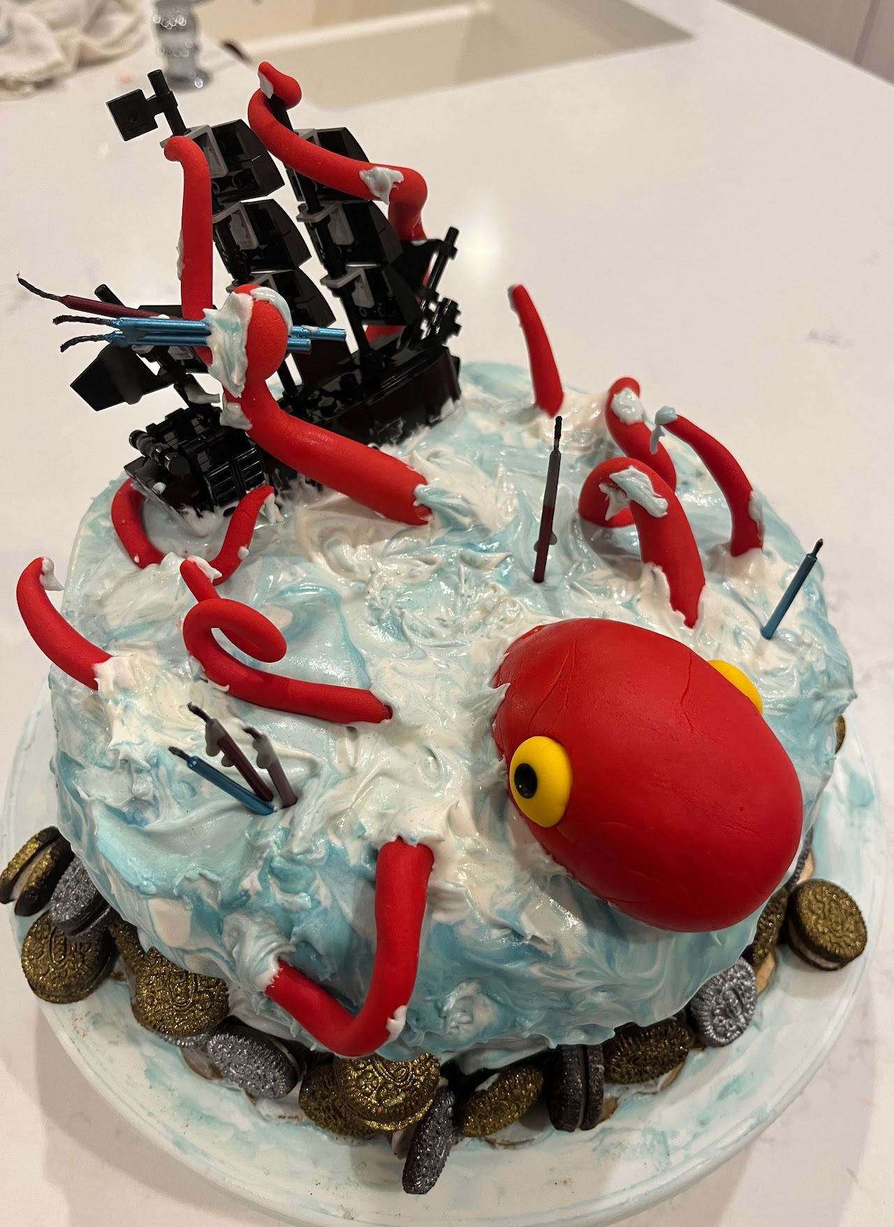 Round cake designed as ocean waves with a giant red fondant kraken pulling down a black pirate ship, gold and silver chocolate coins around the base