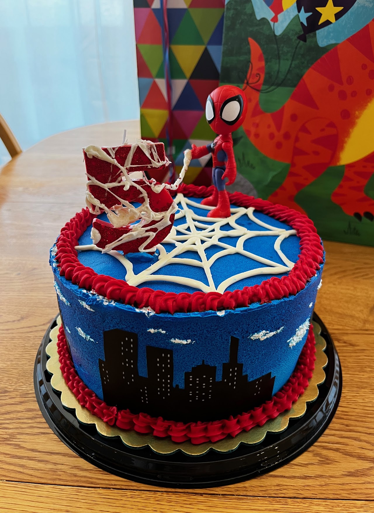 Blue buttercream cake with piped white spiderweb design, a city skyline silhouette, and a Spider-Man figure on top