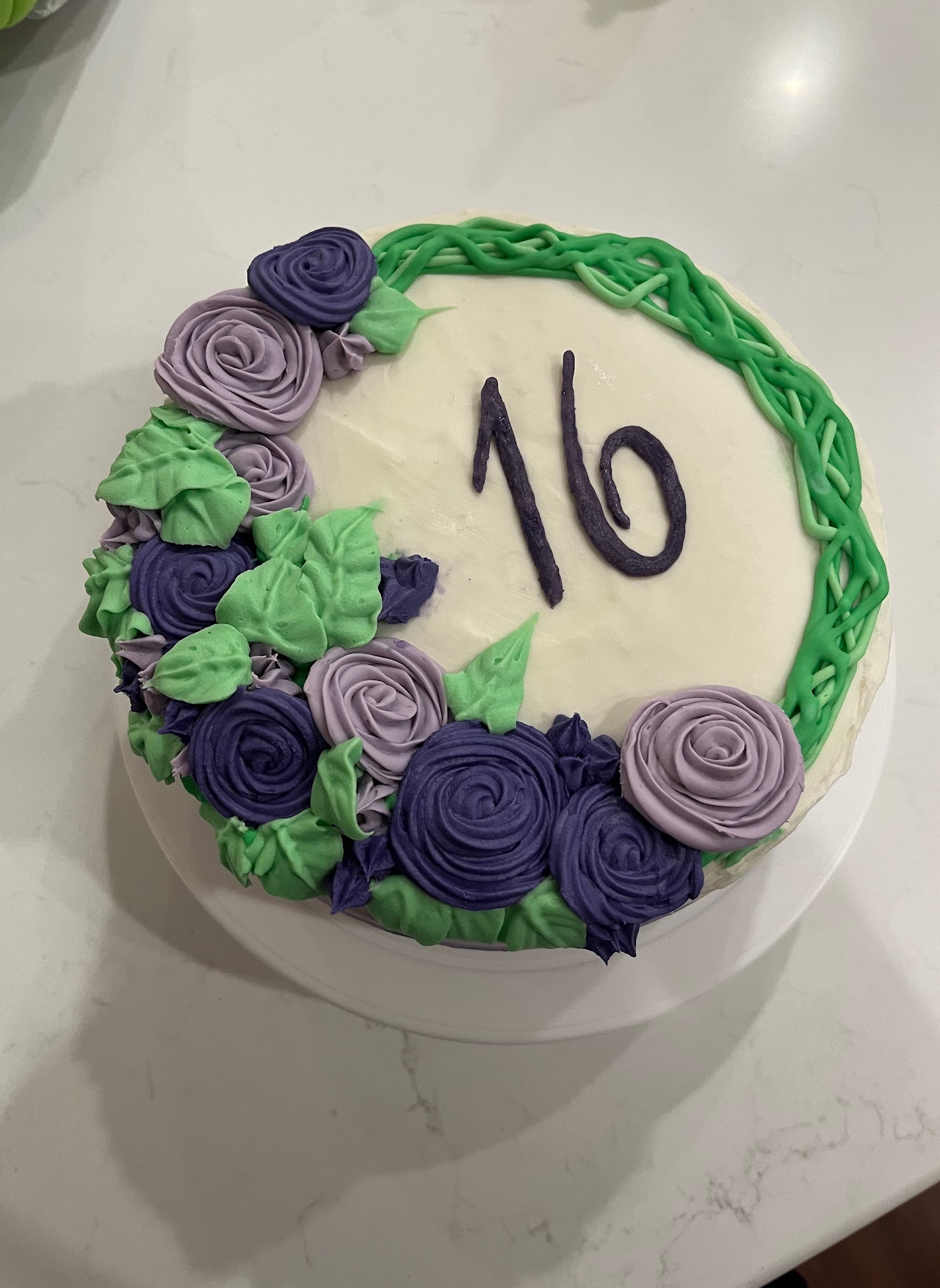White cake with purple and lavender piped buttercream roses, green leaves, and a woven vine border around a hand-piped number 16